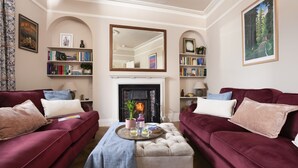 A living room with two red sofas, a central ottoman, a fireplace, and bookshelves with books and decorative items. - Sandy Boobies - Garden, Hot tub, Dog friendly (St Ives)