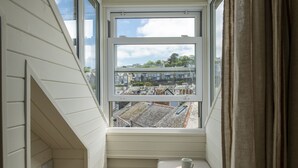 A room with a window overlooking a residential area, a small bench with a cup and book, and a power outlet on the wall. - Sandy Boobies - Garden, Hot tub, Dog friendly (St Ives)
