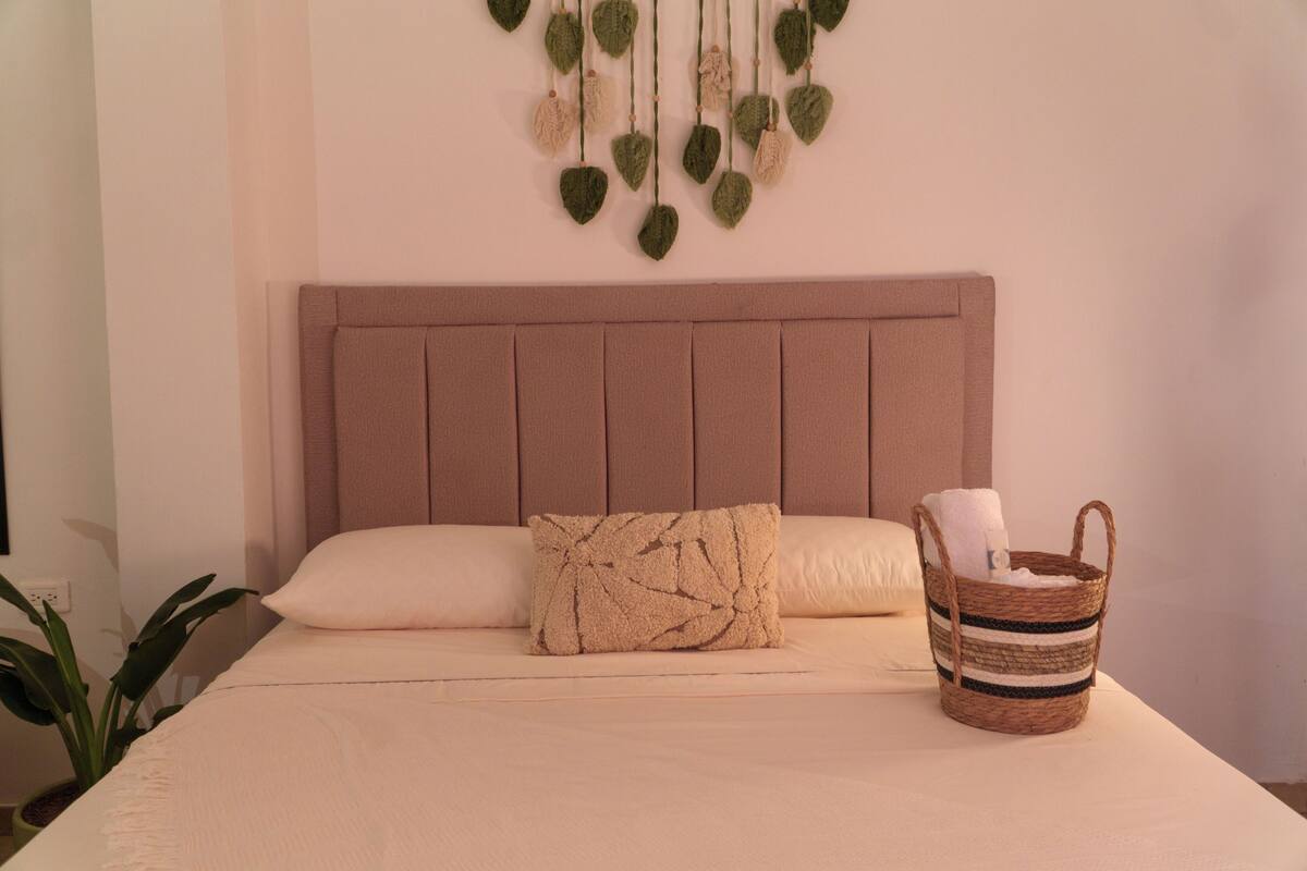 Una cama con cabecera, una almohada, una pequeña cesta tejida y un adorno en la pared.