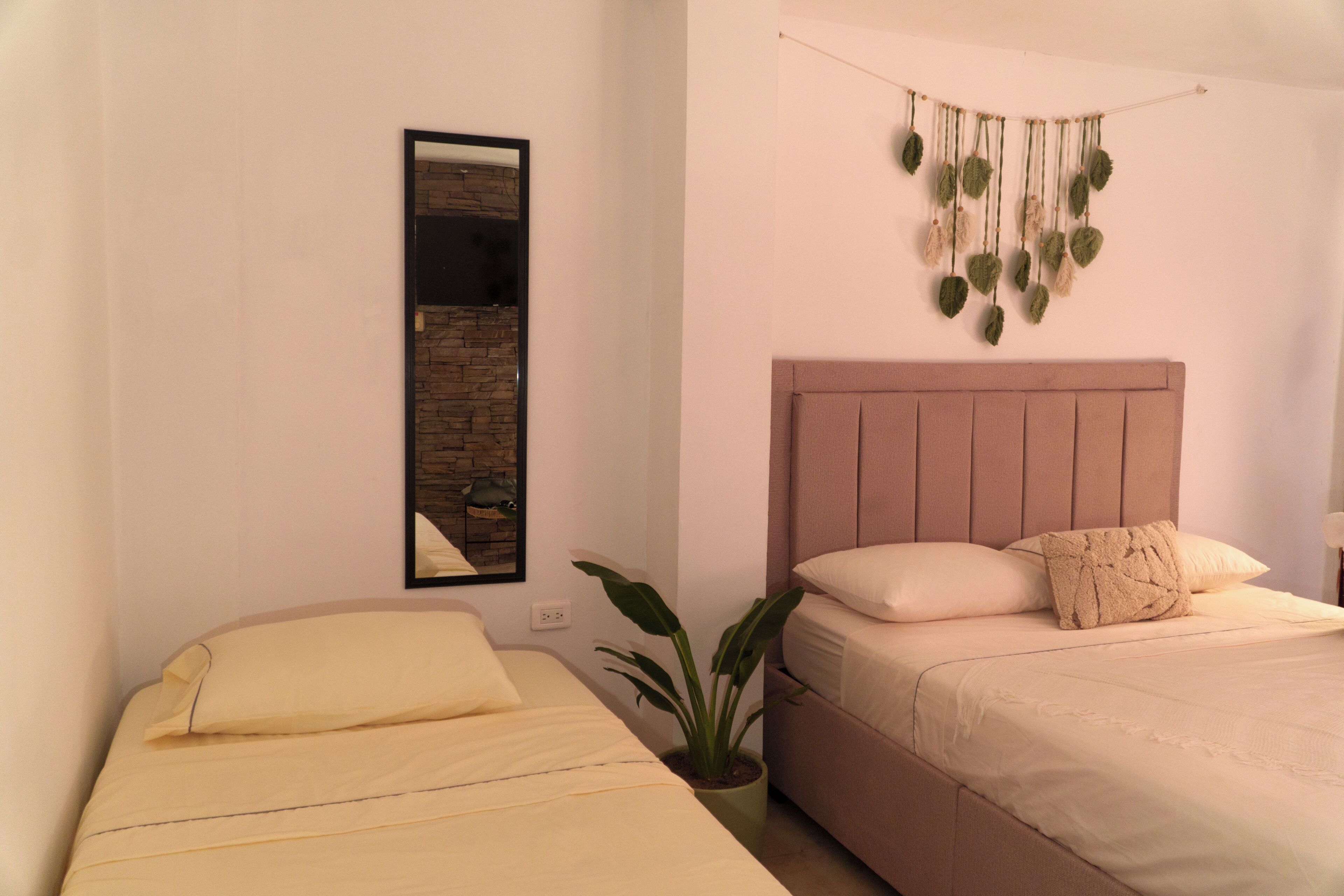Quarto com duas camas, um vaso de planta, um espelho fixado na parede e um adorno pendurado.