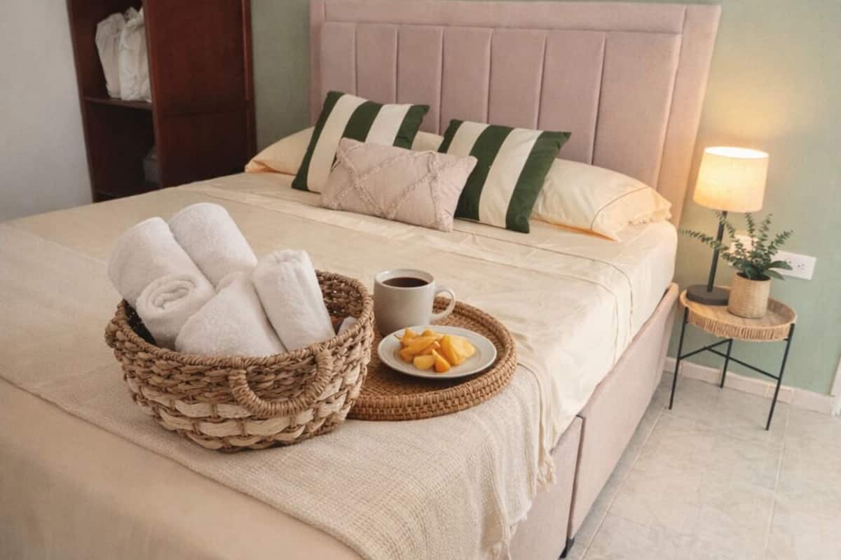 Una cama bien hecha con cabecera, una mesita de noche con lámpara, un cesto de toallas y un plato con fruta.