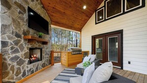 A stone fireplace, a flat-screen TV, a wooden deck, a grill, wicker furniture, and a sliding glass door. - Couples Retreat w/ Hot Tub, Jacuzzi, Fireplaces (Broken Bow)