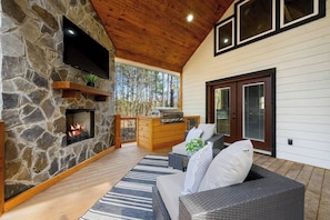Ein Kamin aus Stein, ein Flachbildfernseher, eine Holzterrasse, ein Grill, Korbmöbel und eine Glastür zum Verschieben.