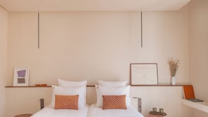 Een tweepersoonsbed met witte beddengoed en twee kussens met oranje motieven, een nachtkastje met een vaas gedroogde bloemen, en een ingelijst certificaat aan de muur.