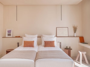 Una cama doble con ropa de cama blanca y dos almohadas anaranjadas estampadas, una mesita de noche con un jarrón de flores secas y un certificado enmarcado en la pared.