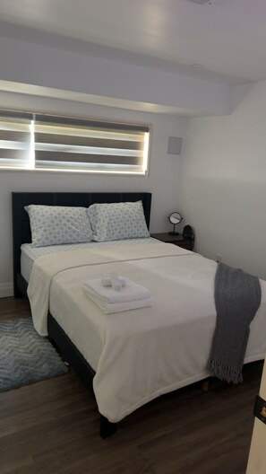 A neatly made bed with a white comforter and pillows, a bedside table with a lamp, and a window with blinds. - 🏡 Modern 2-Bedroom Private Suite Near Niagara Falls | Sleeps 4 | Free Parking (Thorold)