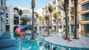 Ein Poolbereich eines Hotels mit Rutsche, Palmen und einem modernen Gebäude mit Balkonen.