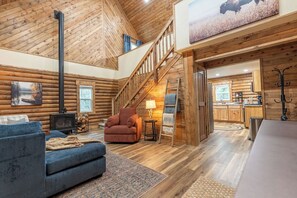 Interior de una cabaña de troncos con chimenea, una escalera, una sala de estar con sofá y sillón, y una zona de cocina.