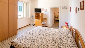 Un dormitorio con cama, armario, televisor, ventilador y una puerta que da a otra habitación.