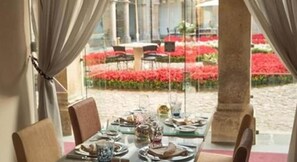 Mesa para dos personas con sillas, vista a un jardín con flores rojas y un pilar de piedra.