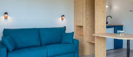 Una habitación moderna con un sofá color turquesa, estanterías de madera, una mesita y una zona de cocina.