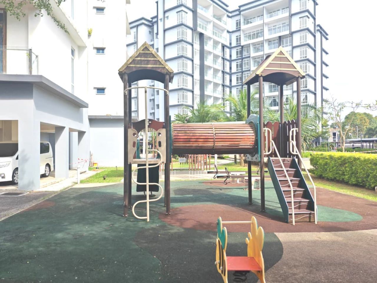 Un parque infantil con estructuras para escalar y un tobogán, rodeado de una zona pavimentada y un edificio al fondo.