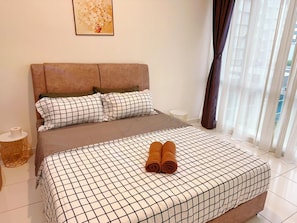 Una cama bien hecha con colcha a cuadros y toallas enrolladas, una mesita de noche con lámpara y una ventana con cortinas traslúcidas.