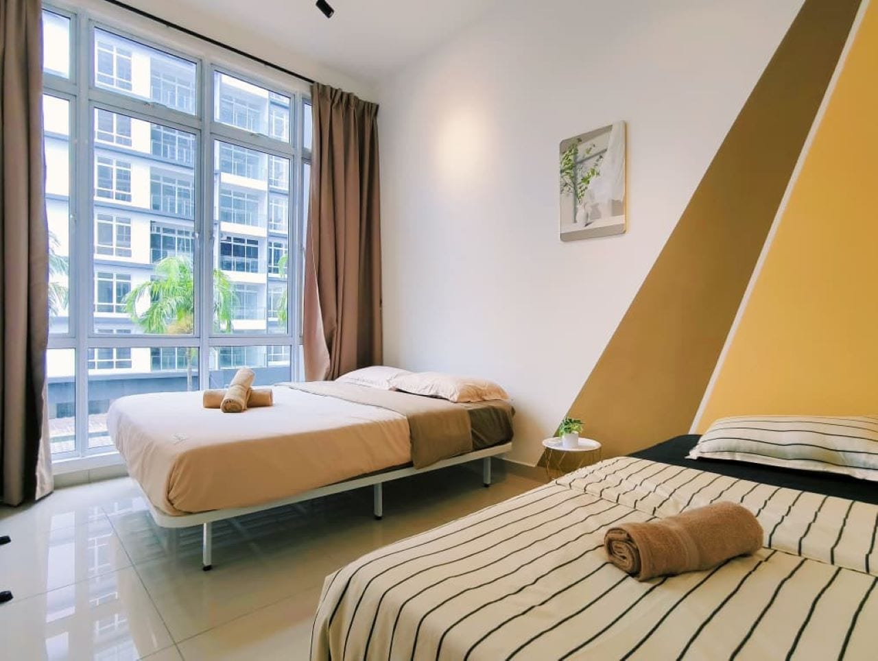 A hotel room with two beds, a large window, a plant on a side table, and a painting on the wall.