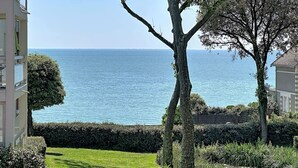 Vista costera con cielo despejado, un camino bordeado de árboles y un edificio con balcones con vista al mar.