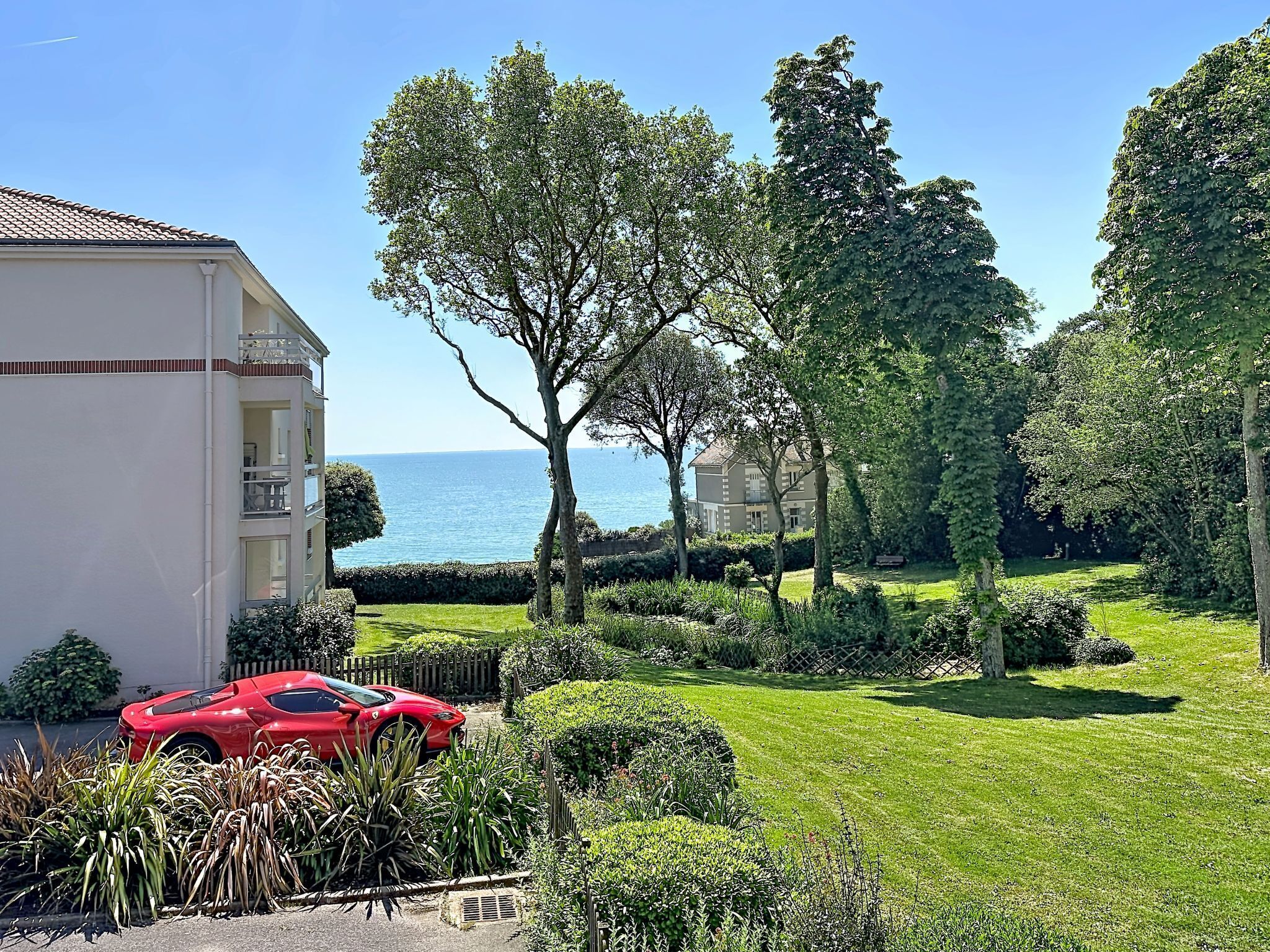 Ein Wohngebiet mit einem roten Sportwagen vor einem Gebäude, einem gepflegten Rasen und Blick auf das Meer.