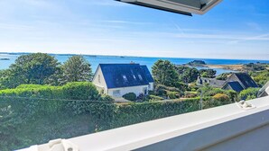 Une vue à travers une fenêtre surmontée d’une lucarne, donnant sur une zone côtière avec des maisons, de la verdure et un ciel bleu dégagé.