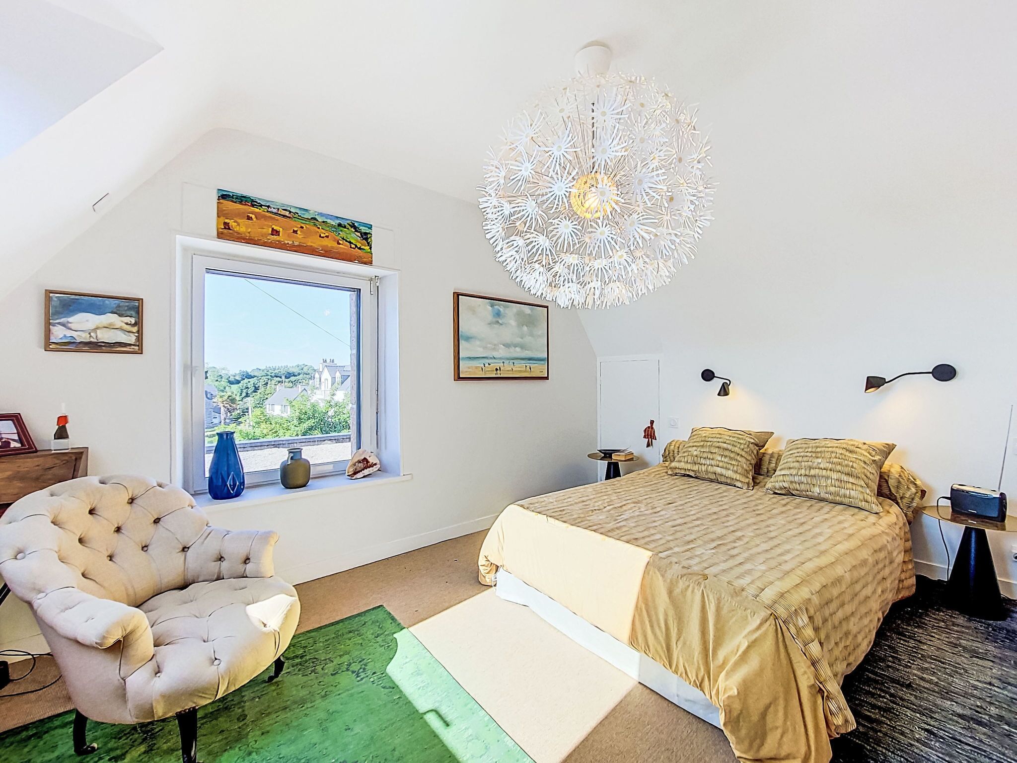 A bedroom with a large bed, a window with a view, a decorative ceiling light, and artwork on the walls.