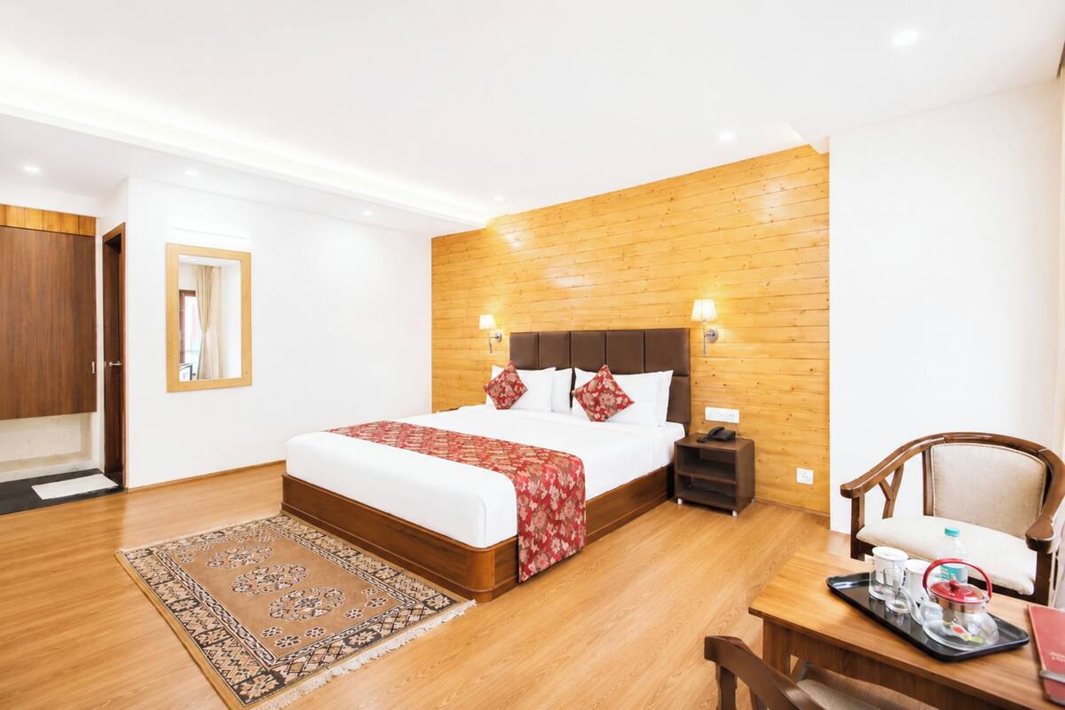 Una habitación de hotel con cabecera de madera, una cama grande, una mesita de noche, una silla, una mesa con bandeja y una alfombra.