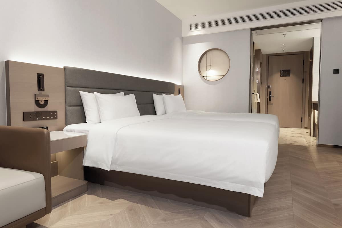 A modern hotel room with a large bed, a bedside table, a chair, a round mirror, and a door leading to another room.