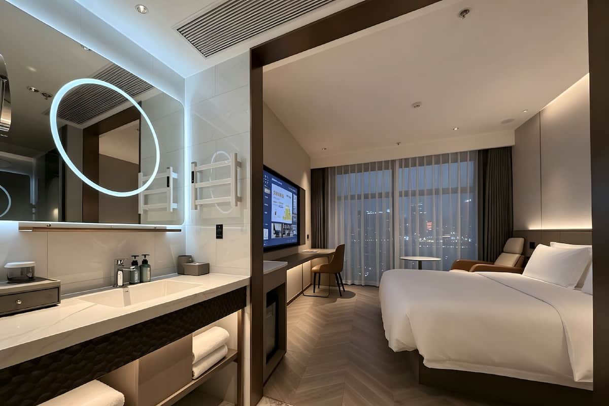 Ein modernes Hotelzimmer mit einem großen Bett, einem Schreibtisch mit Fernseher, einem Badezimmer mit Spiegel und Pflegeprodukten sowie Blick auf die Stadt durch das Fenster.