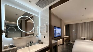 Una habitación de hotel moderna con una cama grande, televisión, baño con espejo grande y lavamanos, y un área de comedor.