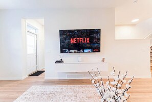 Una sala de estar moderna con un televisor de pantalla plana montado en la pared que muestra el logotipo de Netflix, un mueble bajo blanco y un arreglo floral en el suelo.