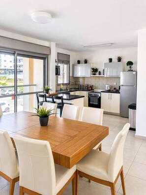 A modern kitchen with white cabinets, a stainless steel refrigerator, and a dining area with a wooden table and white chairs. - Beachfront Apt-Sea View lParking & Mamad (Bat Yam)