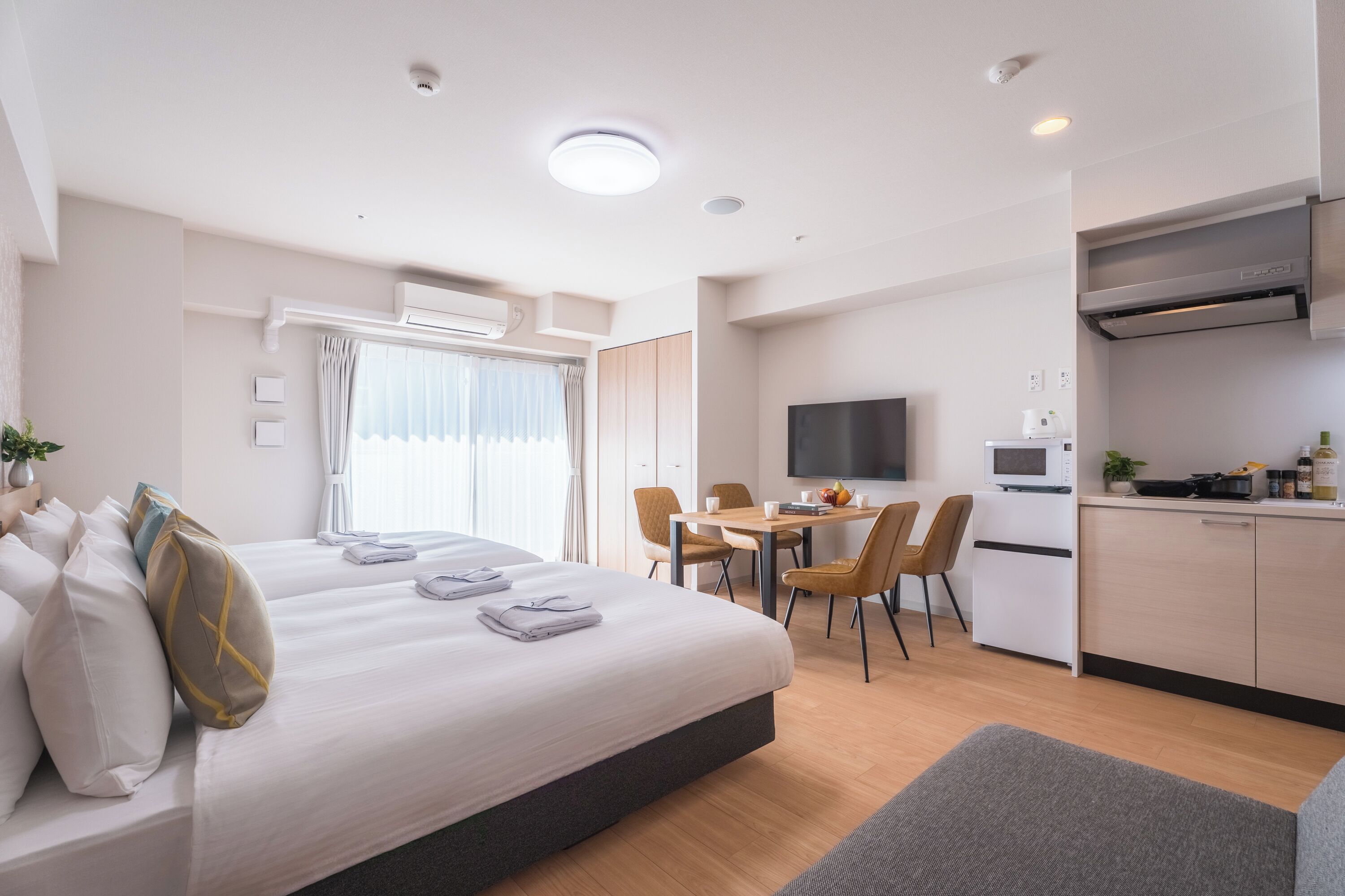 A hotel room with a bed, a dining table with chairs, a kitchen area with a microwave, and a flat-screen TV.