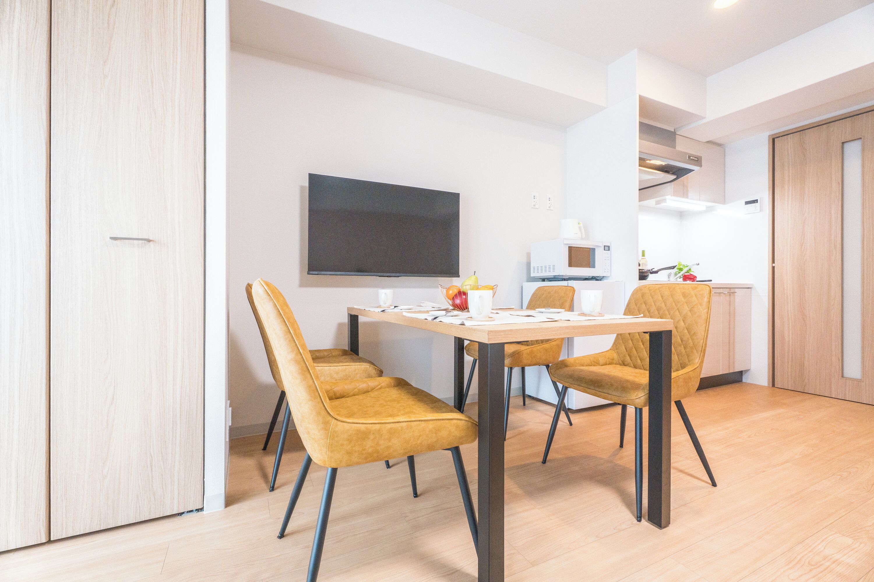 A modern dining area with a table and chairs, a flat-screen TV, and a kitchen in the background.