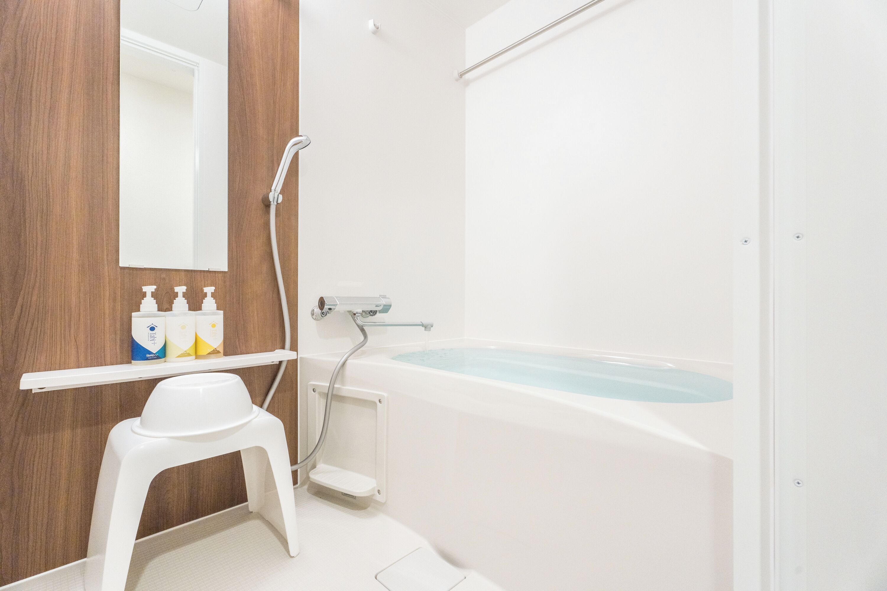 Un baño con una bañera blanca, un taburete blanco, un espejo y un estante con artículos de aseo.
