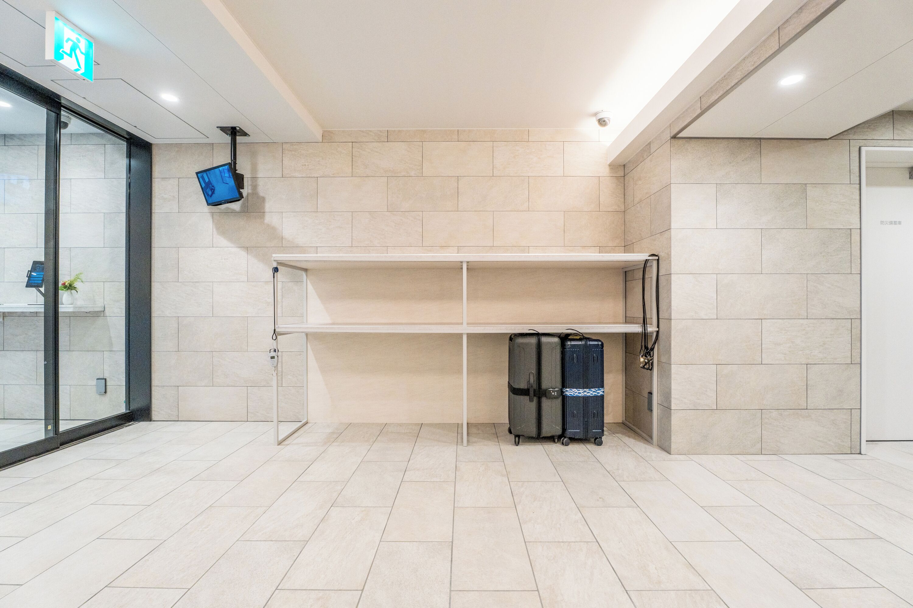 A modern hotel lobby with a tiled floor, a reception desk, a flat-screen TV mounted on the wall, and a glass door entrance.
