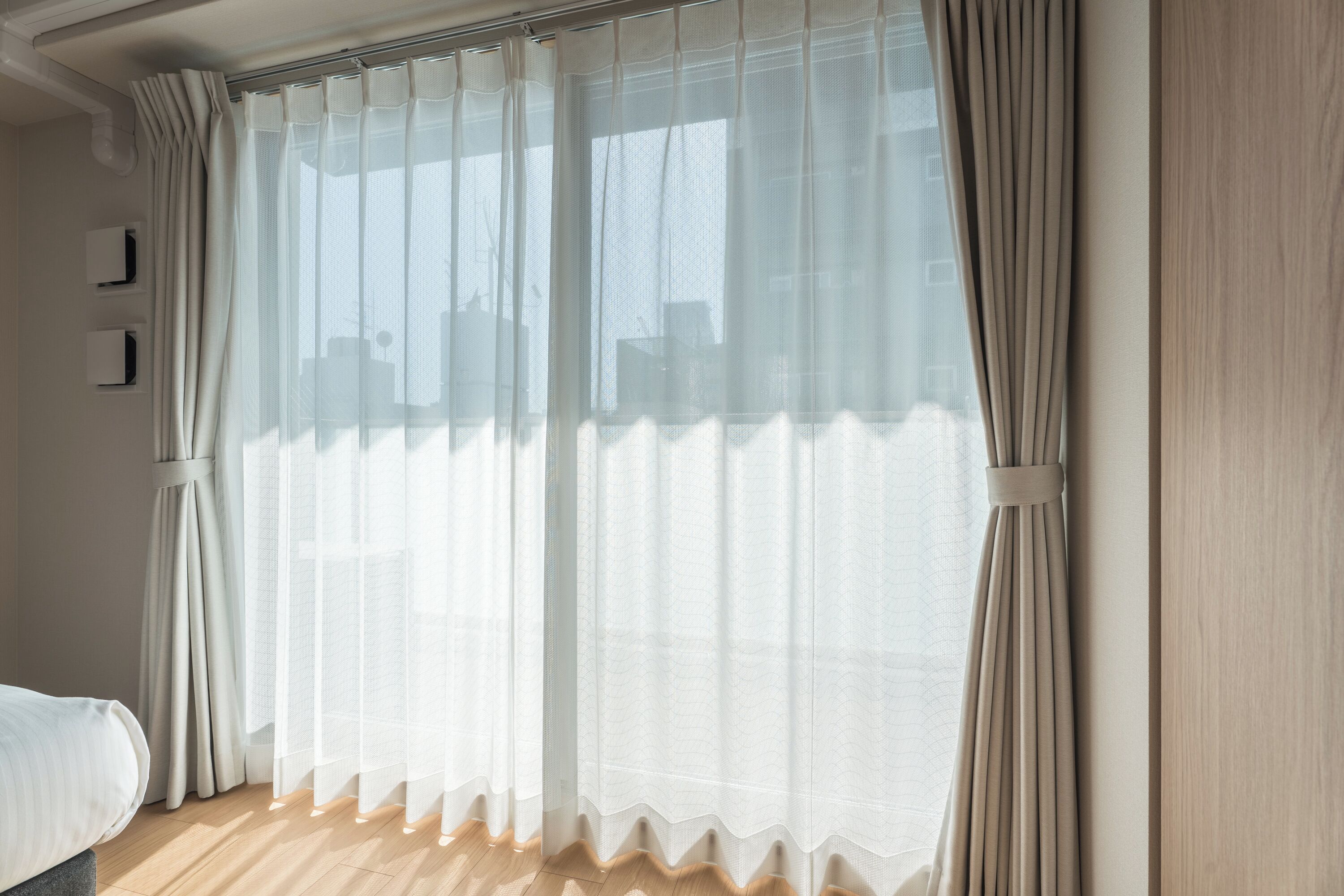 A room with a large window covered by sheer curtains, a bed with a white mattress, and a wooden floor.