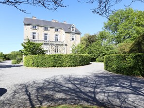 A large stone building with multiple windows and a covered entrance, surrounded by well-maintained hedges and trees.