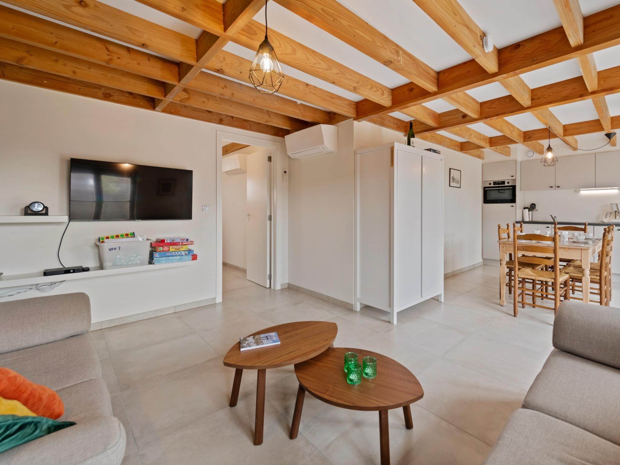 Een moderne woonkamer met een houten plafond, een aan de muur bevestigde televisie, een bank, een salontafel en een eethoek met stoelen.