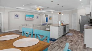 Eine moderne Küche mit Kochinsel, ein Essbereich mit Holztisch und blauen Stühlen sowie ein Wohnzimmer mit grauem Sofa.
