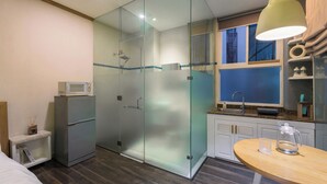 Habitación de hotel moderna con ducha envuelta en vidrio, microondas, refrigerador pequeño, lavamanos y mesa de madera.