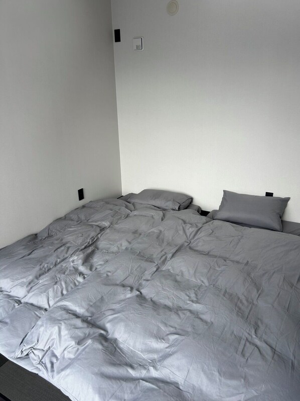 Un lit simple avec une couette grise, un mur blanc et un petit interrupteur sur le mur.