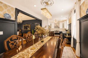 Uma sala de jantar com uma mesa de madeira comprida, cadeiras elaboradas, um lustre e uma cozinha ao fundo.