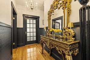 Um corredor com porta preta, espelho dourado ornamentado e lustre.