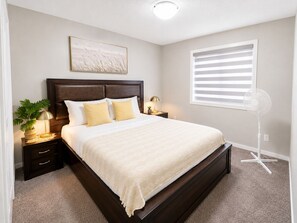 A neatly made bed with a dark headboard, a bedside lamp, and a floor fan. - Stay Cool! -Modern 3BR Home Near YYC with New Central AC (Calgary)