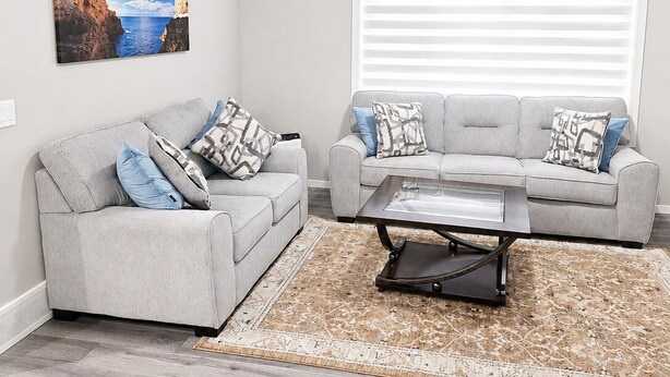 A living room with two grey sofas, a coffee table, a patterned rug, and a large painting of a coastal scene. - Stay Cool! -Modern 3BR Home Near YYC with New Central AC (Calgary)