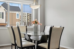 A dining area with a glass table, black chairs, a vase with flowers, and a view of buildings through large windows. - Stay Cool! -Modern 3BR Home Near YYC with New Central AC (Calgary)