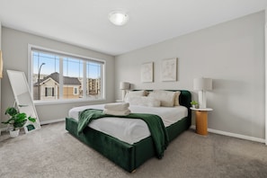 A bedroom with a large window, a bed with a green headboard, two bedside lamps, and a small side table with a plant. - Stay Cool! -Modern 3BR Home Near YYC with New Central AC (Calgary)