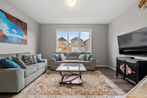 A living room with a gray sofa, a glass coffee table, a patterned rug, a television, and a painting of a coastal scene. - Stay Cool! -Modern 3BR Home Near YYC with New Central AC (Calgary)