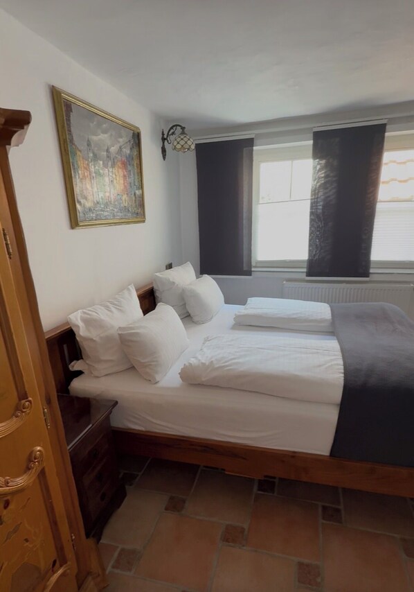 Un dormitorio con cama, almohadas, una mesita de noche, cómoda, ventana con cortinas y un cuadro en la pared.