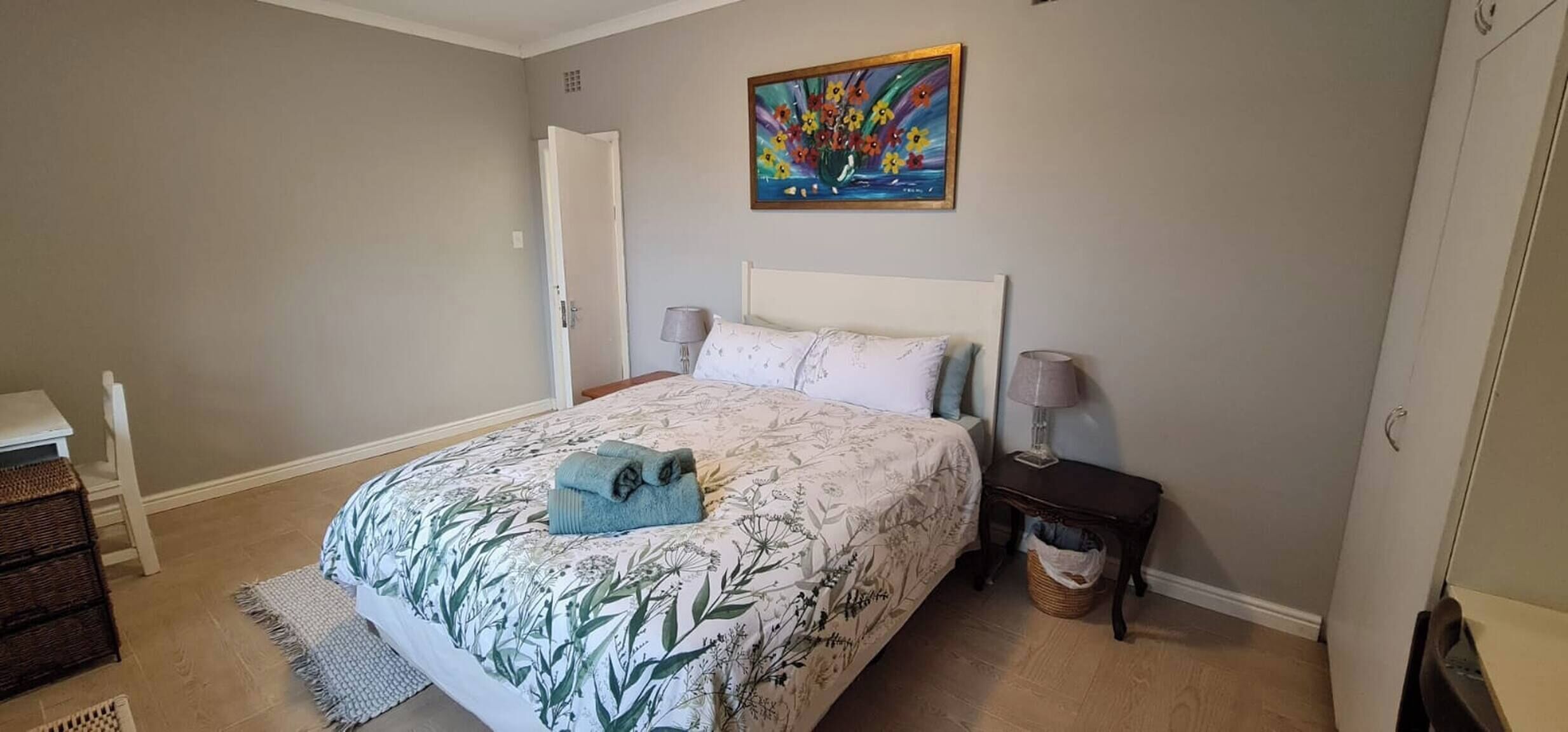 A bedroom with a bed, bedside tables, a lamp, a painting, a door, and a small wicker basket.