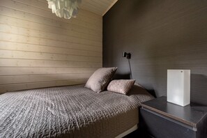 A bedroom with a wooden sloped ceiling, a bed with a textured blanket, two pillows, a bedside table with a lamp, and a white cabinet. - Villa Lumi 4 | Cozy Arctic Retreat in Ylläs (Ylläsjärvi)