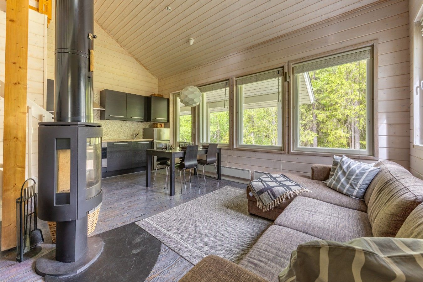 A cozy cabin interior with a wood-burning stove, a dining area, a sofa, and large windows.