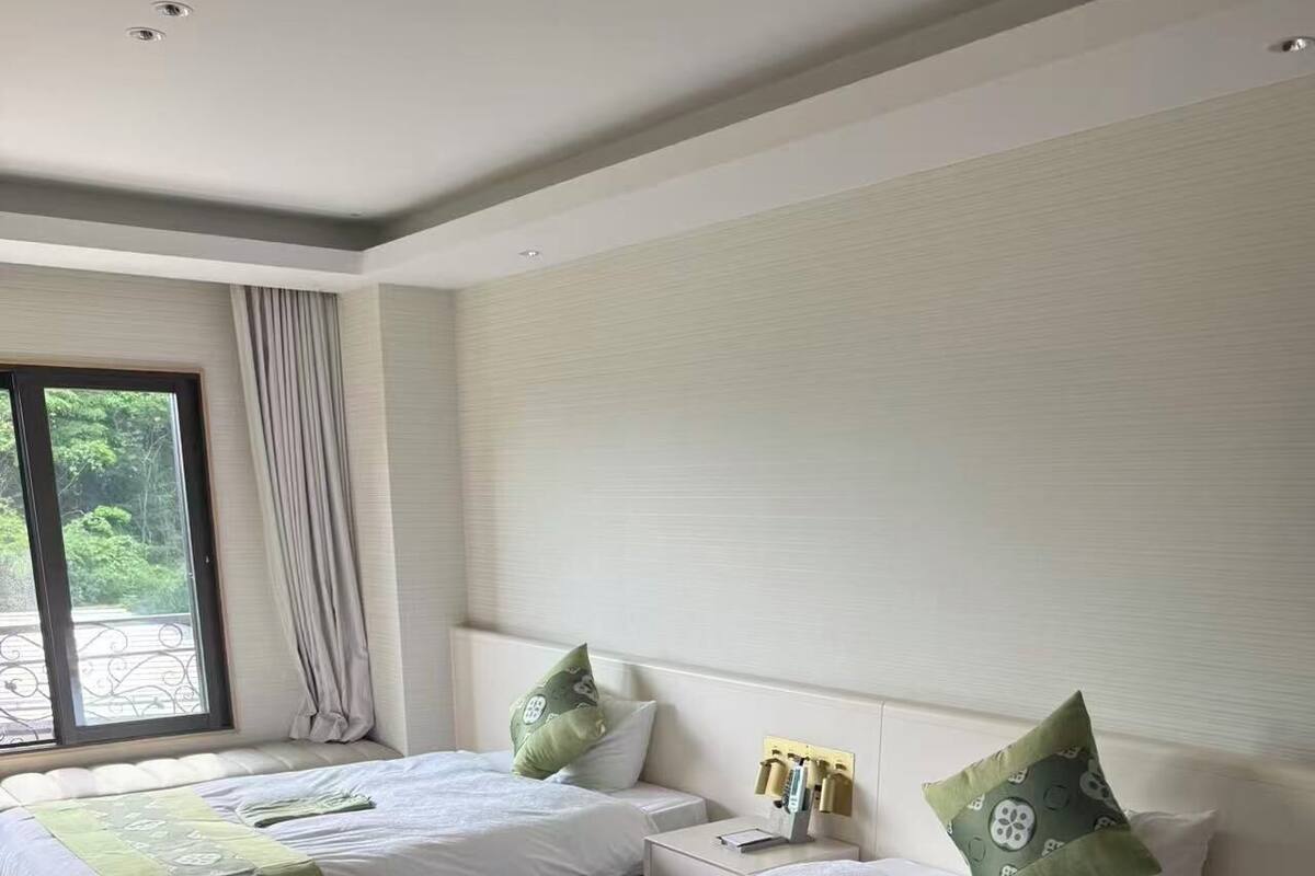 A hotel room with two beds, a bedside table with a lamp, a window with curtains, and a view of greenery outside.